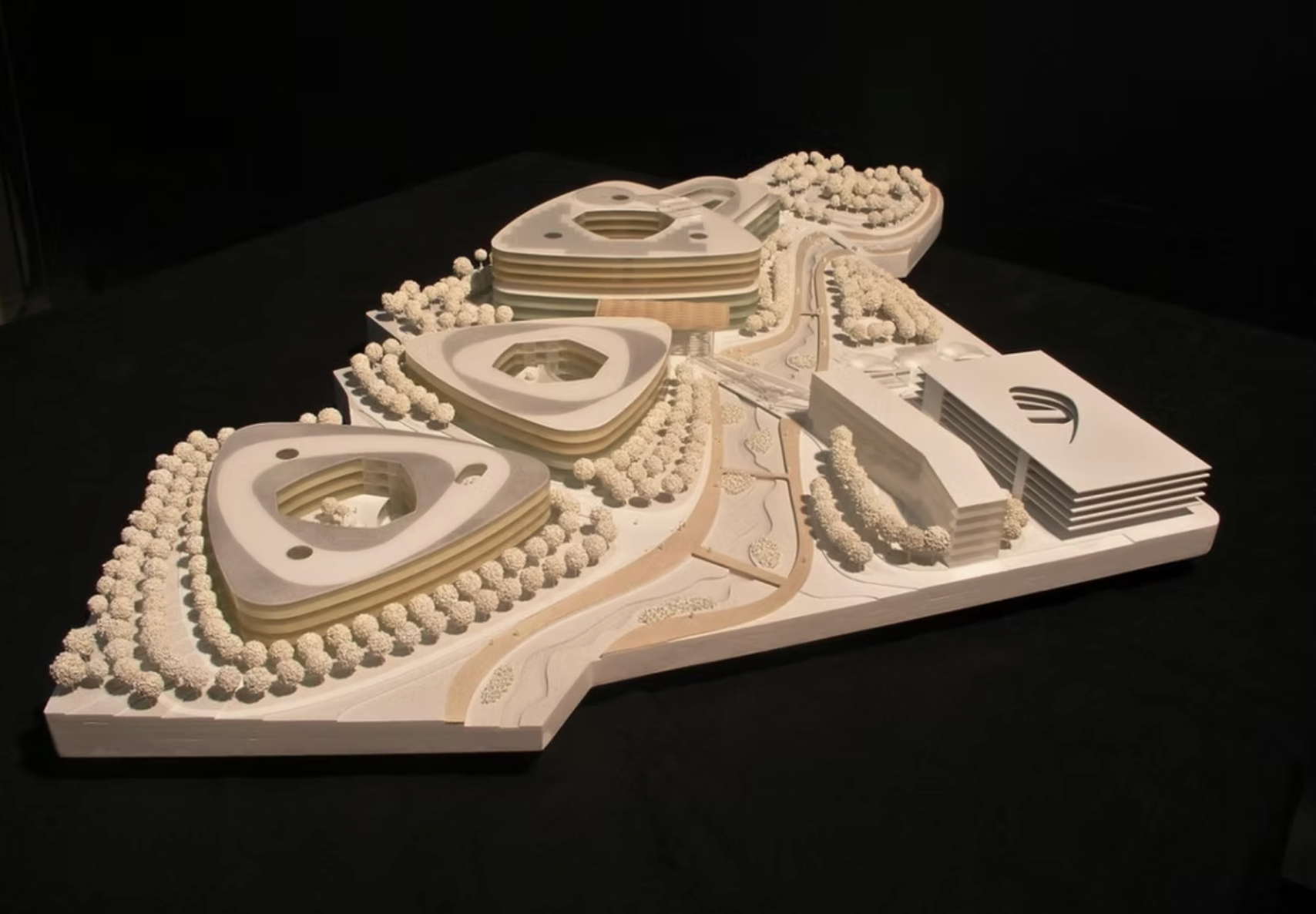 Maquette en bois d’un projet urbain futuriste avec trois édifices triangulaires à étages formant des patios centraux, un bâtiment rectangulaire adjacent, des routes sinueuses et de petites sculptures d’arbres dispersées sur le site.
