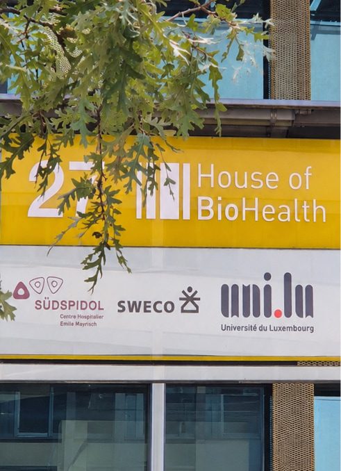 Façade du bâtiment avec enseigne jaune « 2 House of BioHealth » à Luxembourg, accompagnée des logos « SÜDSPIDOL Centre Hospitalier Emile Mayrisch », « SWECO » et « Université du Luxembourg », avec un rameau de chêne en premier plan.